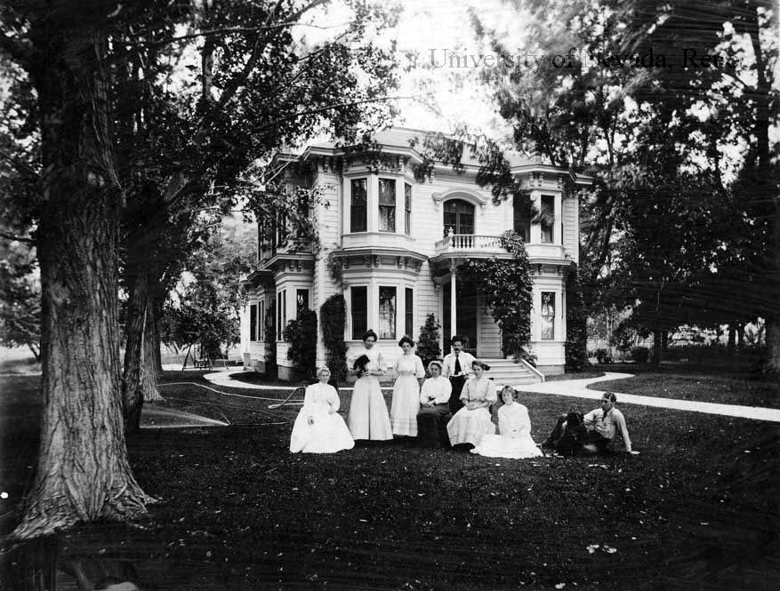 This black and white image shows six women in white dresses, some holding black fans, and two men in the yard of the Evans home.